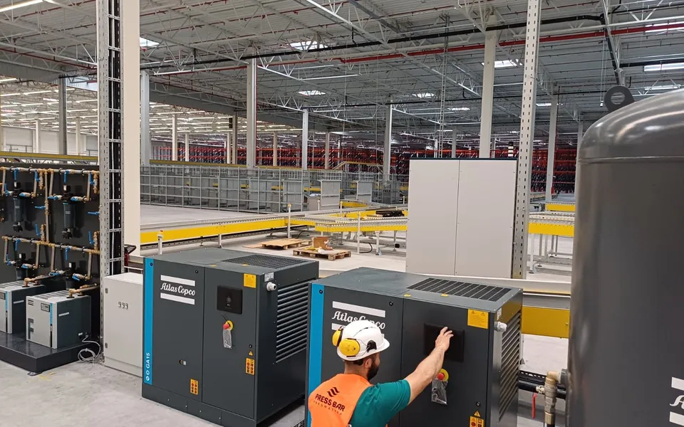 Serwisant Press Bar uruchamia sprężarkę śrubową Atlas Copco w hali przemysłowej; obok kolejne sprężarki i osuszacze